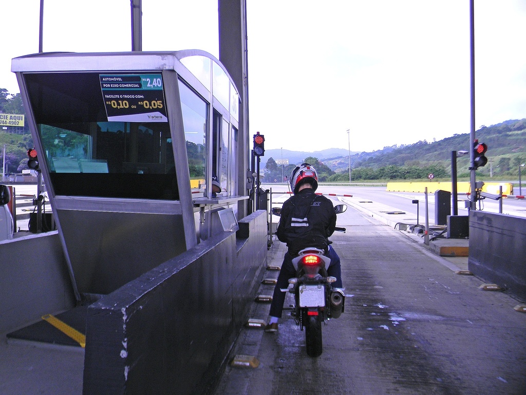 Na imagem um motociclista vestindo jaqueta preta e capacete branco com detalhes vermelhos está parado em uma cabine de pedágio em uma rodovia brasileira. A cabine, situada à esquerda, exibe uma placa informativa com valores em Reais e solicita moedas para facilitar o troco. O motociclista aguarda a liberação da cancela, enquanto ao fundo observa-se a continuidade da estrada cercada por vegetação e colinas sob um céu claro.