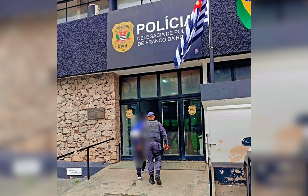 Um policial militar fardado e usando boina azul é visto de costas enquanto conduz um homem algemado, que veste uma jaqueta azul e branca, subindo uma rampa de concreto em direção à entrada de vidro de uma delegacia. A fachada do edifício combina uma parede texturizada escura na parte superior, onde está fixada uma placa preta com o brasão da Polícia Civil do Estado de São Paulo e os dizeres POLÍCIA - DELEGACIA DE POLÍCIA DE FRANCO DA ROCHA, com uma parede lateral feita de pedras rústicas claras. À direita da entrada, hasteadas em mastros, aparecem as bandeiras do estado de São Paulo e do Brasil, completando o cenário institucional.