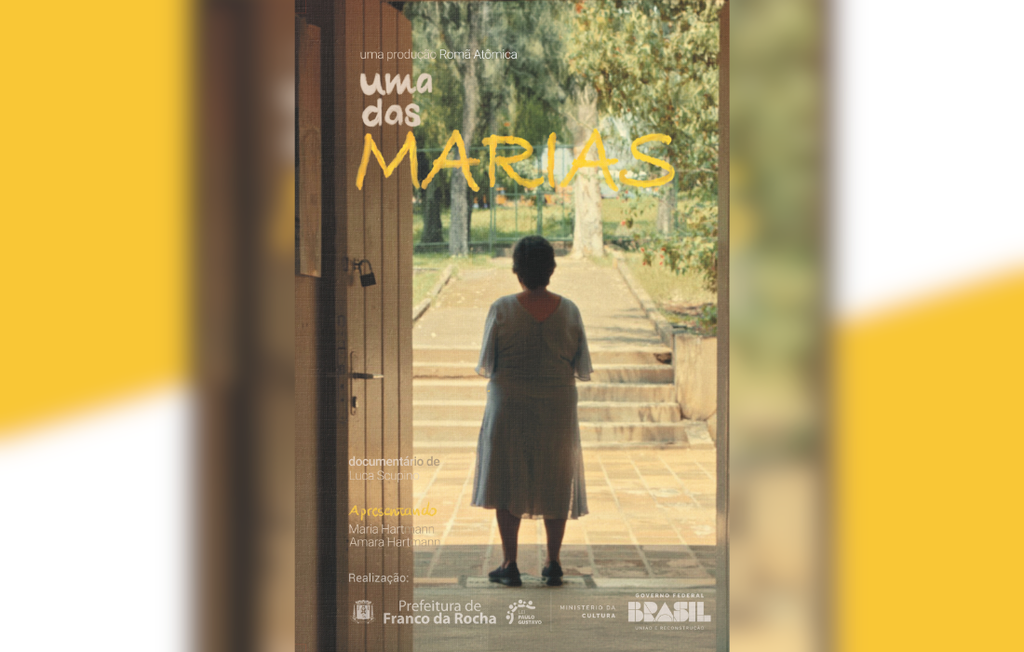 Imagem mostra uma mulher de costas, em pé diante de uma porta aberta, olhando para um jardim com árvores e degraus de pedra. A luz natural ilumina o ambiente, criando um clima contemplativo. Sobre a imagem, o título “Uma das Marias” aparece em letras brancas e amarelas.