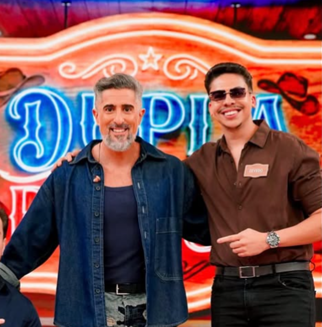 Dois homens posam lado a lado diante de um letreiro iluminado com a palavra “DUPLA”. À esquerda, o apresentador Marcos Mion, um homem de cabelos grisalhos e barba que veste camisa jeans azul escura sobre camiseta marinho. À direita, outro homem usa óculos escuros, camisa marrom com crachá escrito “ZEVEDO” e relógio no pulso. O fundo é colorido e decorado com elementos de estilo country, como chapéus de cowboy e estrelas, sugerindo ambiente festivo ou de entretenimento.