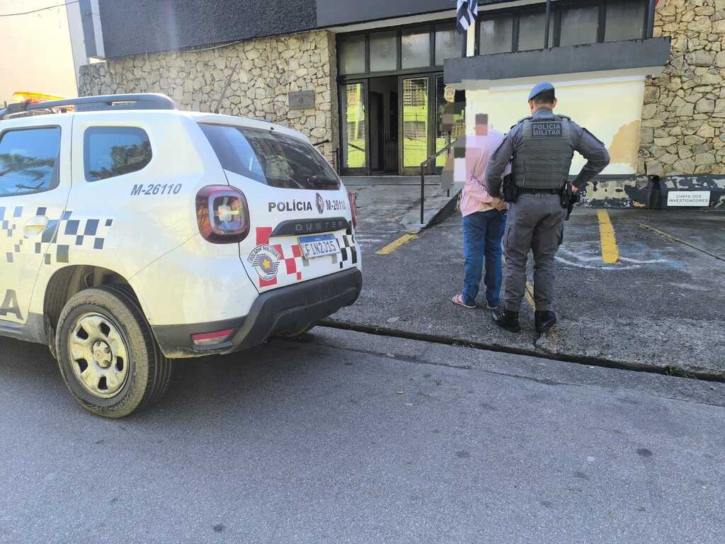 Um policial militar fardado e de boina azul, visto de costas, escolta um homem algemado com as mãos para trás, que veste camisa rosa clara e calça jeans, no pátio externo de uma delegacia com paredes de pedras rústicas. À esquerda, em primeiro plano, está estacionada a traseira de uma viatura branca modelo Renault Duster da Polícia Militar de São Paulo, com grafismos quadriculados em cinza, identificação 