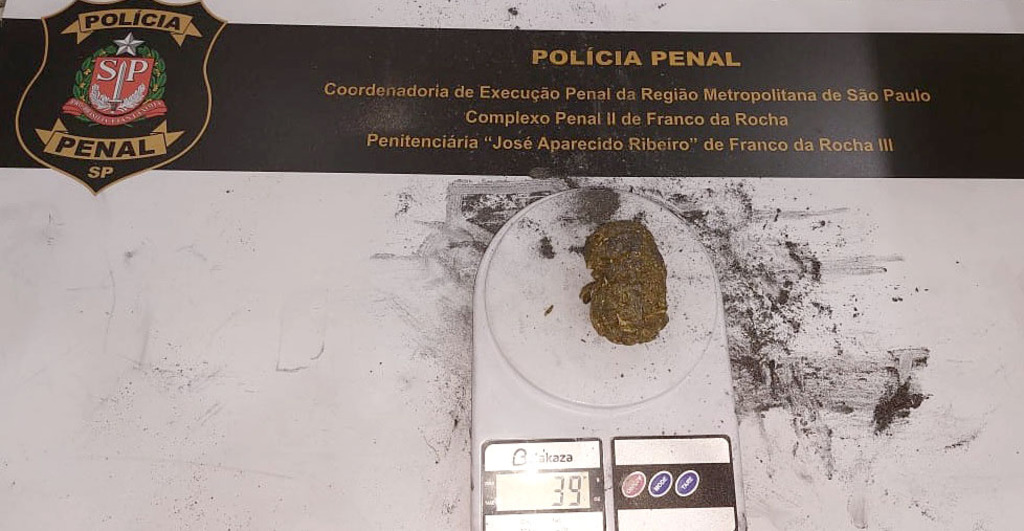 A imagem mostra uma pequena porção de uma substância esverdeada e prensada, semelhante à maconha, posicionada sobre uma balança digital de precisão branca da marca Akaza, que indica o número 39 em seu visor. Ao fundo, sobre uma superfície clara com resíduos de pó escuro espalhados, há um banner oficial da Polícia Penal do Estado de São Paulo, contendo o brasão da instituição à esquerda e os textos 