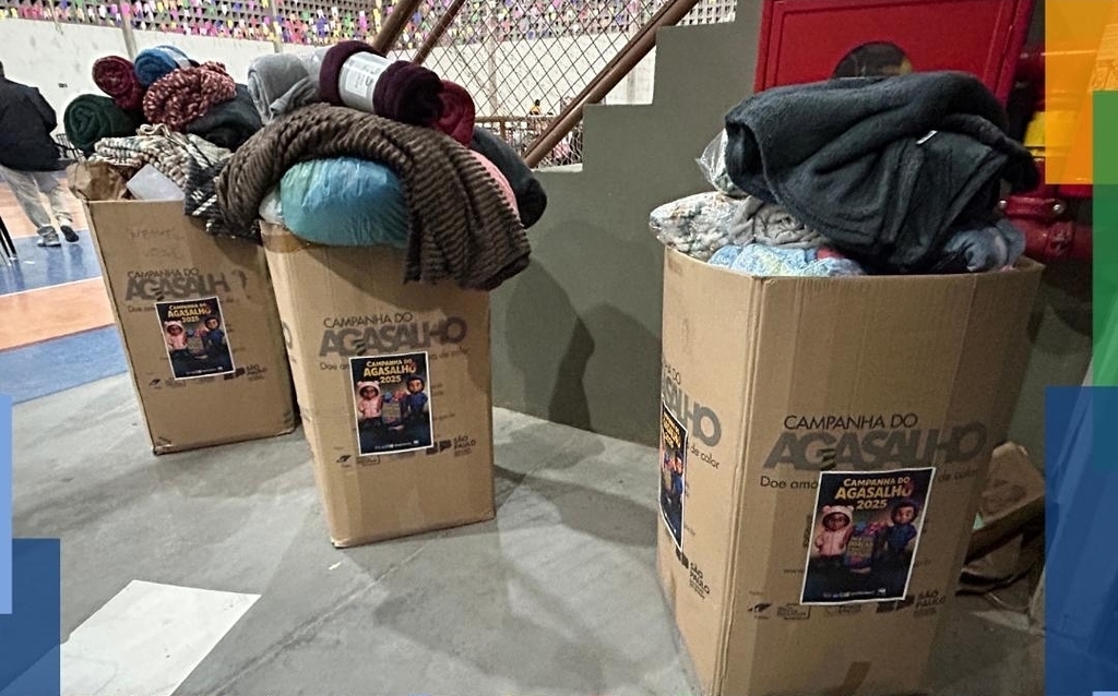A imagem mostra três grandes caixas de papelão repletas de agasalhos e cobertores doados, posicionadas em um ambiente interno que parece ser um ginásio. Cada caixa exibe um cartaz oficial da 