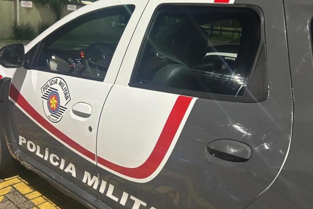 A lateral de uma viatura da Polícia Militar do Estado de São Paulo é vista em ângulo diagonal, exibindo sua característica pintura nas cores branco, cinza escuro e detalhes em vermelho. Na porta dianteira, destaca-se o brasão da corporação com a estrela de cinco pontas centralizada, enquanto a inscrição 