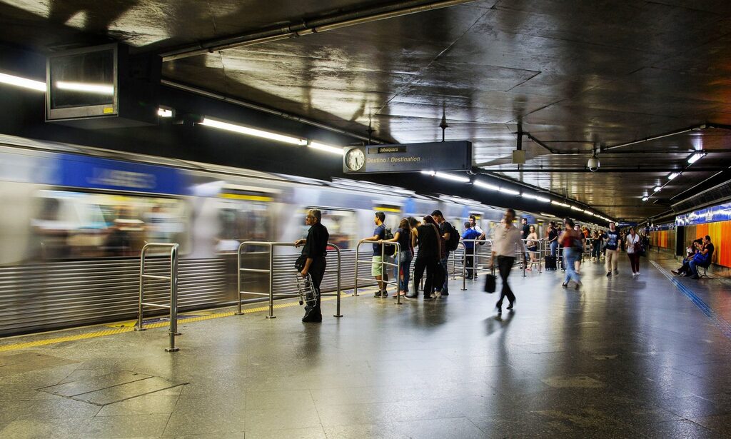 Uma fotografia em plano médio e ângulo levemente baixo captura o interior de uma estação de metrô movimentada, onde o movimento é enfatizado pelo rastro borrado de um trem prateado com detalhes azuis partindo à esquerda. No centro da composição, diversos passageiros aguardam na plataforma de piso cinza polido e reflexivo; um homem de preto observa os trilhos em primeiro plano, enquanto outros pedestres caminham ou esperam próximos às barras de proteção metálicas. A iluminação é composta por fileiras de lâmpadas fluorescentes no teto de concreto escuro, que criam linhas de perspectiva em direção ao fundo da estação, onde se veem paredes com faixas horizontais em azul e laranja, além de placas informativas suspensas que indicam o destino 