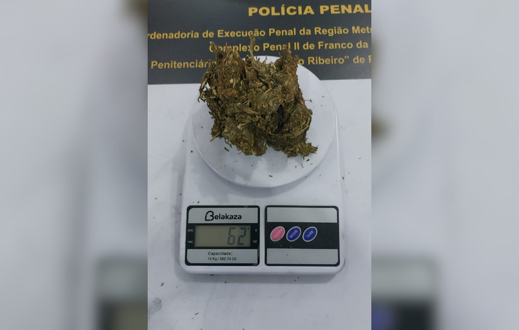 Uma balança digital branca da marca Belakaza está posicionada no centro da imagem sobre uma superfície clara, exibindo o número 62 no visor digital ao lado da unidade de medida em gramas (g). Sobre o prato redondo da balança, há uma porção volumosa de substância vegetal prensada e seca em tons de verde-escuro e marrom, assemelhando-se a maconha.