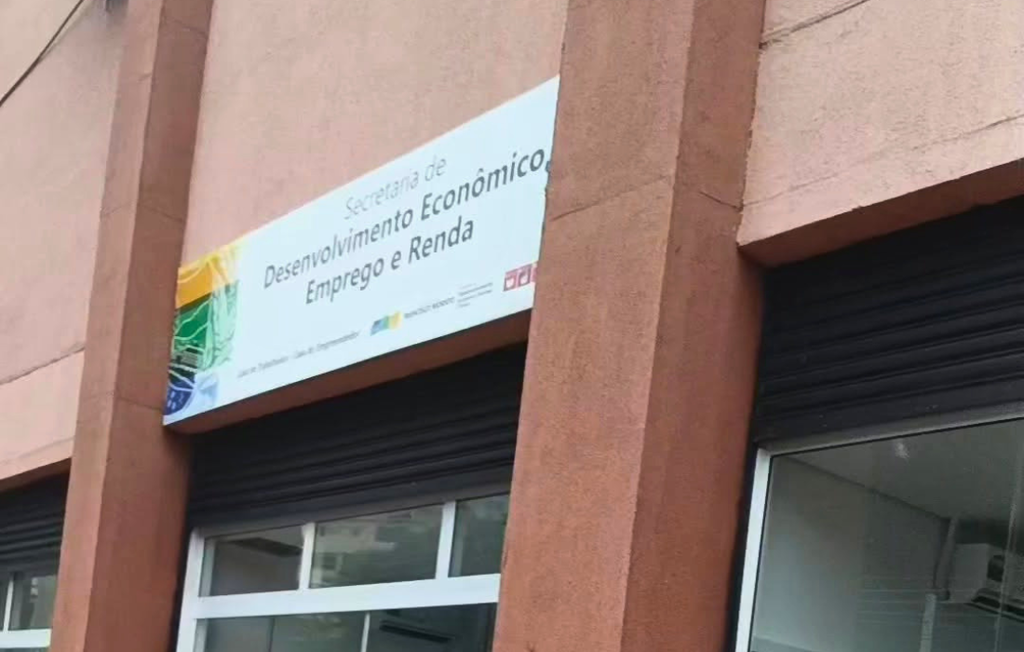 Fotografia em ângulo contra-plongée da fachada de um edifício de alvenaria pintado em tom terracota, destacando uma placa retangular branca fixada acima de uma porta de enrolar preta. A placa exibe o logotipo oficial do governo à esquerda e o texto centralizado em letras escuras: 