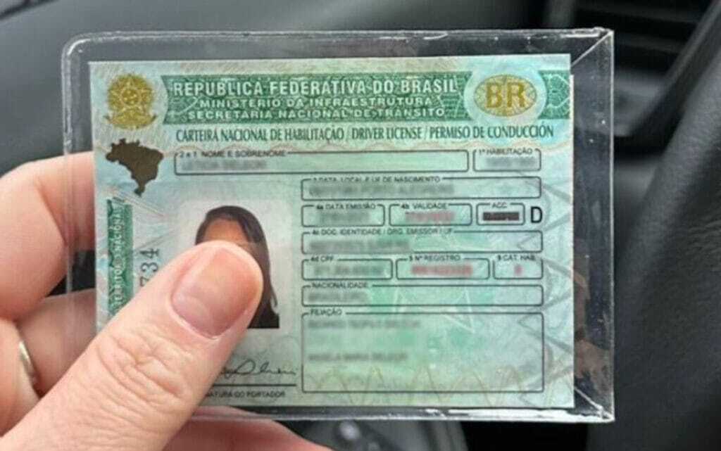 Uma mão segura, dentro de um protetor plástico transparente, uma Carteira Nacional de Habilitação (CNH) brasileira com o modelo atualizado, apresentando detalhes em verde e amarelo, o brasão da República e o mapa do Brasil no canto superior esquerdo. O documento exibe a foto de uma mulher parcialmente coberta pelo polegar de quem o segura, além de campos preenchidos com informações pessoais, como nome, data de nascimento, filiação e o número de registro, destacando-se a categoria 