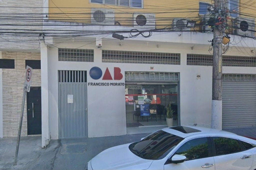 A imagem mostra a fachada branca da OAB de Francisco Morato, com seu logotipo colorido e uma entrada de vidro. Em primeiro plano, destaca-se a frente de um carro branco e, acima, fiações elétricas e aparelhos de ar-condicionado.