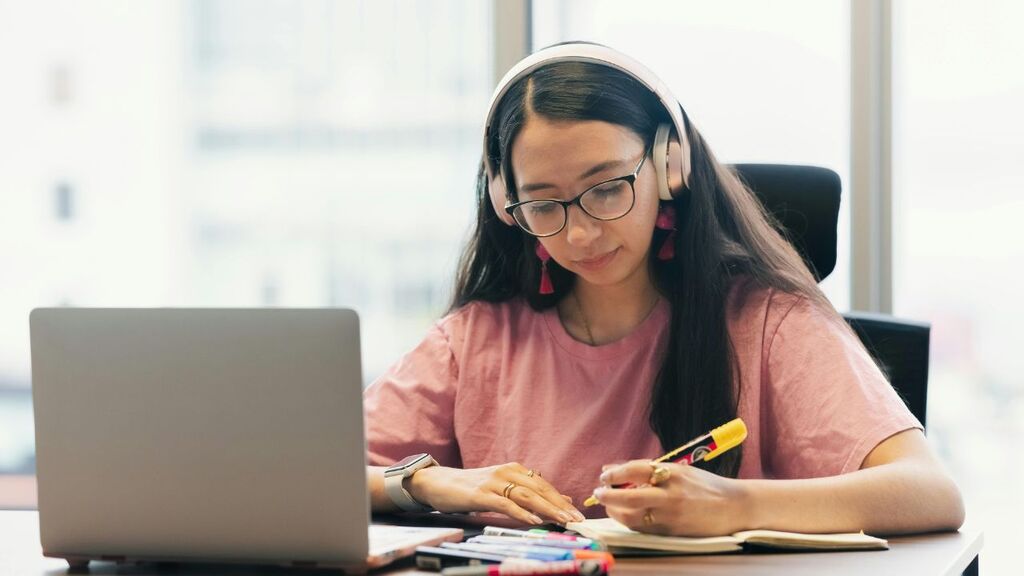 A imagem mostra uma jovem sentada a uma mesa, profundamente concentrada no seu trabalho. Tem cabelos longos e escuros, usa óculos redondos e auscultadores de ouvido cor-de-rosa. Veste uma camisa cor-de-rosa e um relógio branco no pulso esquerdo. À sua frente, encontra-se um computador portátil prateado, que está aberto e ligeiramente voltado para a direita. A jovem segura um marcador amarelo na mão direita, parecendo prestes a fazer anotações num caderno aberto, que está sobre a mesa. A mão esquerda está apoiada no caderno.