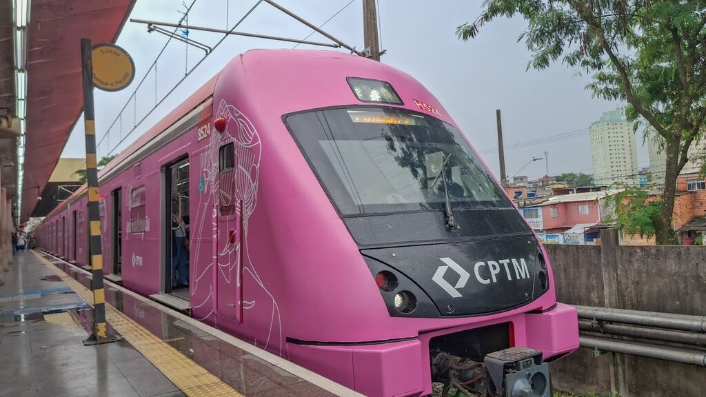 Um trem da CPTM com a lataria inteiramente adesivada na cor rosa e grafismos brancos está parado em uma plataforma de estação sob um céu nublado. A frente do trem exibe o logotipo da companhia em destaque sobre uma base preta, enquanto a lateral apresenta as portas abertas para embarque e desembarque, com a estrutura da plataforma e elementos urbanos ao fundo completando o cenário.