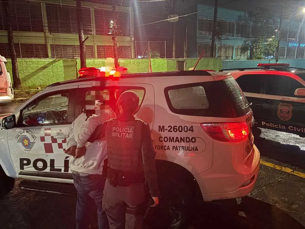 A imagem mostra uma cena noturna em que um policial militar fardado, de costas para a câmera, segura uma pessoa algemada com as mãos para trás ao lado de uma viatura branca da Polícia Militar do Estado de São Paulo. A viatura exibe as inscrições 