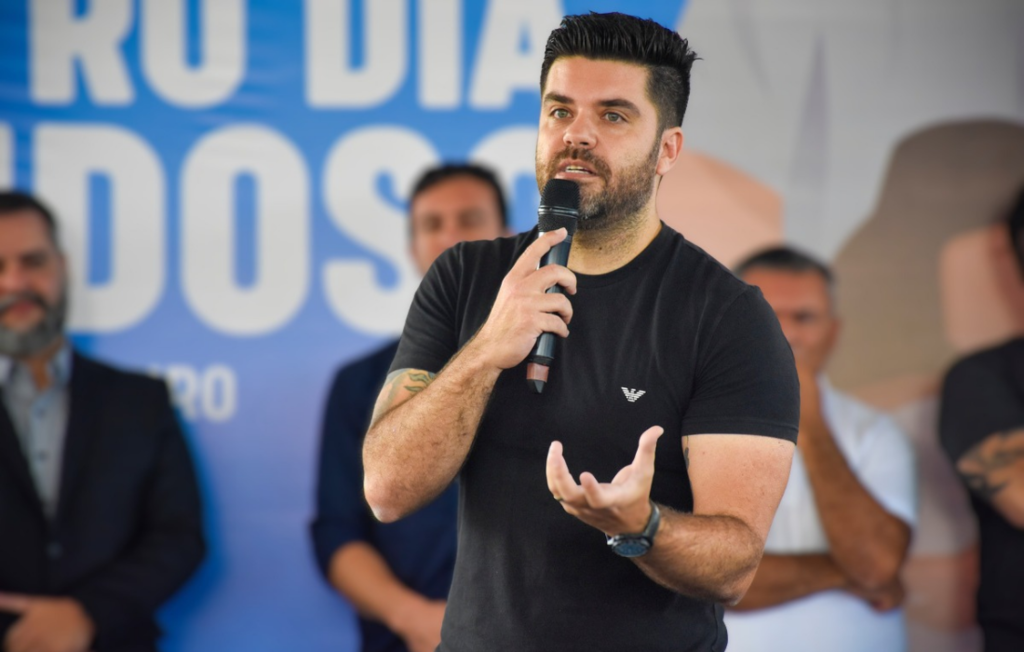 A imagem mostra um homem jovem, de pele clara e cabelos escuros curtos, falando ao microfone enquanto gesticula com a mão esquerda em um evento público. Ele veste uma camiseta preta com um pequeno logotipo de marca no peito e possui tatuagens visíveis no braço direito. Ao fundo, desfocados, estão outros homens e um painel azul com letras brancas grandes, sugerindo um ambiente de discurso ou conferência política.