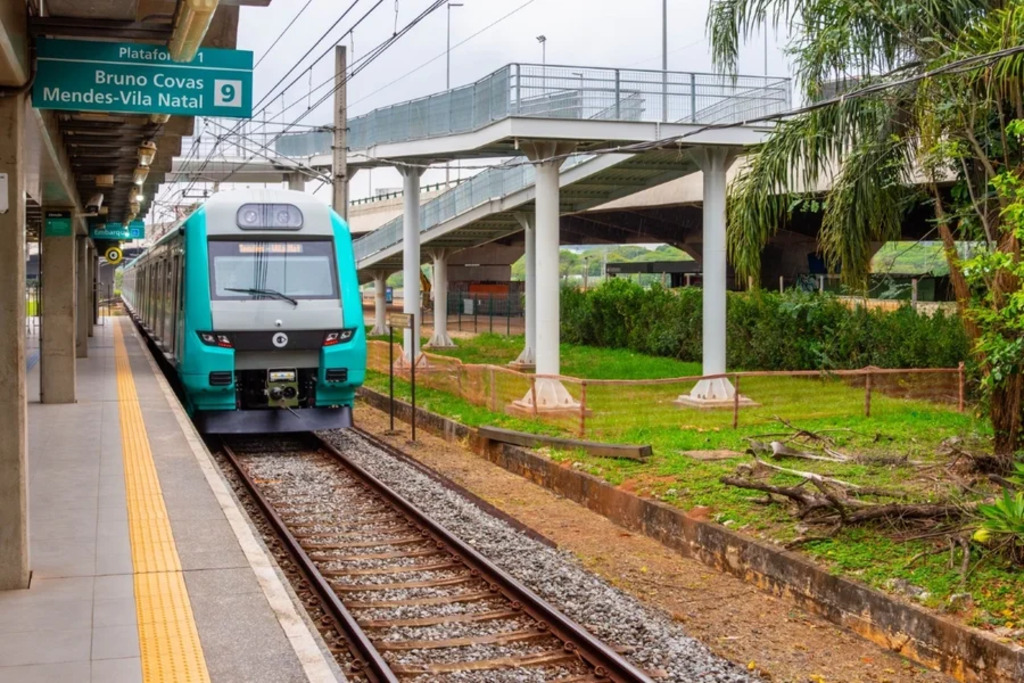 Trem moderno nas cores turquesa e cinza chegando à plataforma de uma estação. O piso possui faixa tátil amarela ao longo da borda e, acima, há uma placa verde indicando “Plataforma 1” e “Bruno Covas Mendes-Vila Natal”, com o número 9 em destaque. A estrutura da estação é limpa e organizada, com passarela elevada sustentada por colunas brancas cruzando sobre os trilhos. Ao redor, há vegetação com arbustos e palmeiras, compondo o cenário urbano integrado a áreas verdes.
