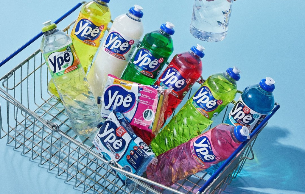 Uma fotografia publicitária tirada de um ângulo superior mostra um carrinho de compras metálico preenchido com diversos produtos de limpeza da marca Ypê sobre um fundo azul claro uniforme. Dentro do carrinho, destacam-se várias garrafas plásticas de detergente líquido em cores vibrantes, como amarelo, verde, vermelho, rosa, azul e uma versão branca de coco, todas organizadas de forma inclinada. Além dos líquidos, há embalagens de esponjas de limpeza posicionadas ao centro, enquanto uma mão (parcialmente visível no topo direito) segura uma garrafa de detergente transparente, sugerindo o ato de abastecer o carrinho. A cena possui uma iluminação clara e direta, ressaltando o brilho das embalagens e a variedade do portfólio da marca.