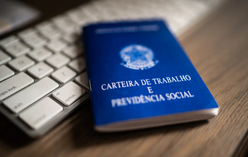 Fotografia em ângulo diagonal e com efeito de desfoque mostrando uma Carteira de Trabalho e Previdência Social (CTPS) brasileira azul, em primeiro plano e levemente inclinada sobre uma mesa de madeira escura. O documento exibe o brasão da República Federativa do Brasil em branco e as inscrições 