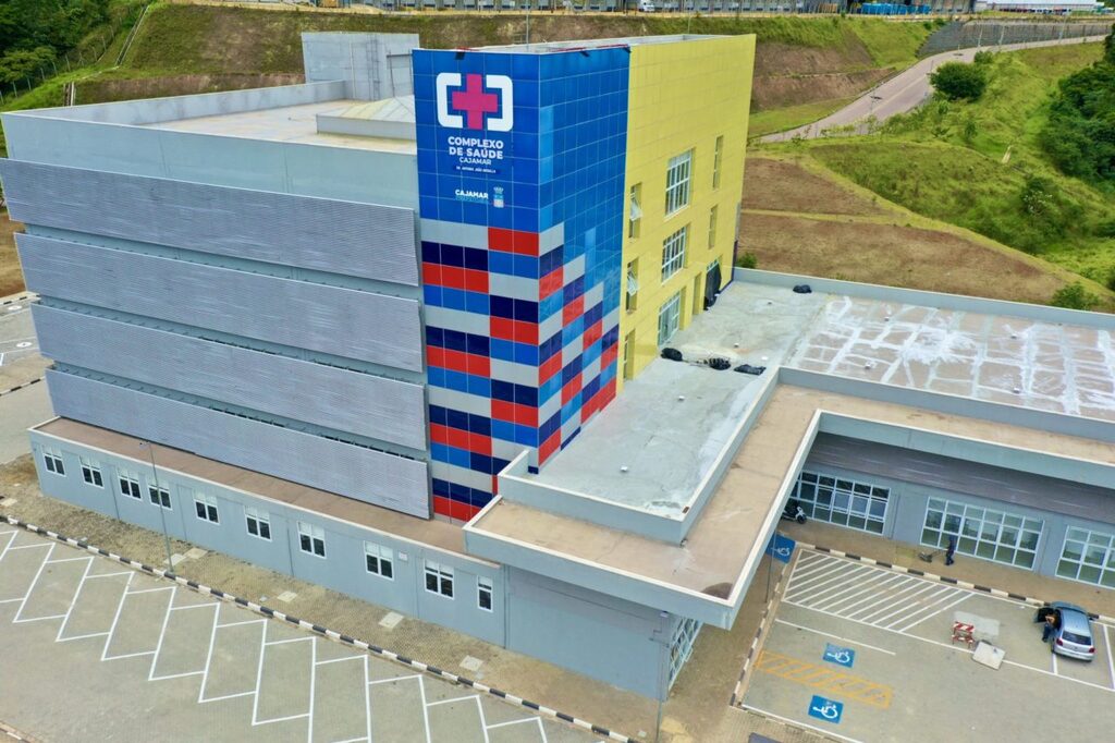 Imagem aérea do Complexo de Saúde de Cajamar mostra um edifício moderno de grande porte, com fachada colorida em tons de azul, vermelho e branco e identificação visual na torre central; o local conta com ampla área externa, estacionamento sinalizado com vagas acessíveis e está situado em uma região com áreas verdes e vias de acesso ao redor