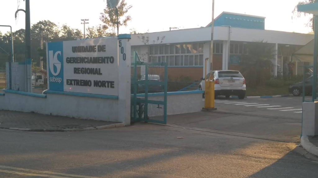 Entrada da unidade da Sabesp identificada como “Unidade de Gerenciamento Regional Extremo Norte”. O portão azul está parcialmente aberto, e ao lado há uma placa branca com o logotipo azul da Sabesp e o número 500. Dentro do terreno, vê-se um prédio de um andar com janelas amplas e um carro branco estacionado. A iluminação indica fim de tarde, com sombras suaves e árvores ao fundo.