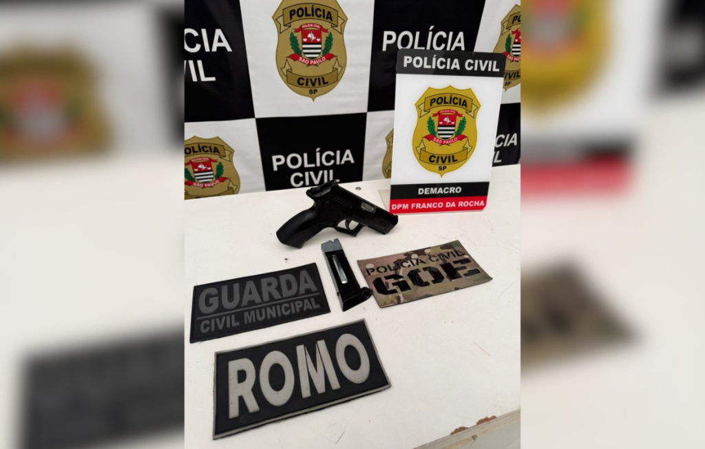 A imagem mostra uma pistola preta e seu carregador, aparentemente de ar comprimido (co2), dispostos sobre uma mesa branca ao lado de três tarjetas de identificação policial com as inscrições 