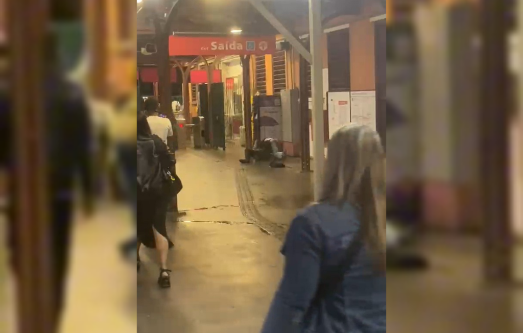 A imagem mostra o interior de uma estação de trem iluminada à noite, com algumas pessoas transitando pelo local. Em primeiro plano, uma mulher de costas e cabelos compridos veste uma jaqueta jeans azul. Mais à frente, no corredor de piso claro, uma placa vermelha suspensa indica 