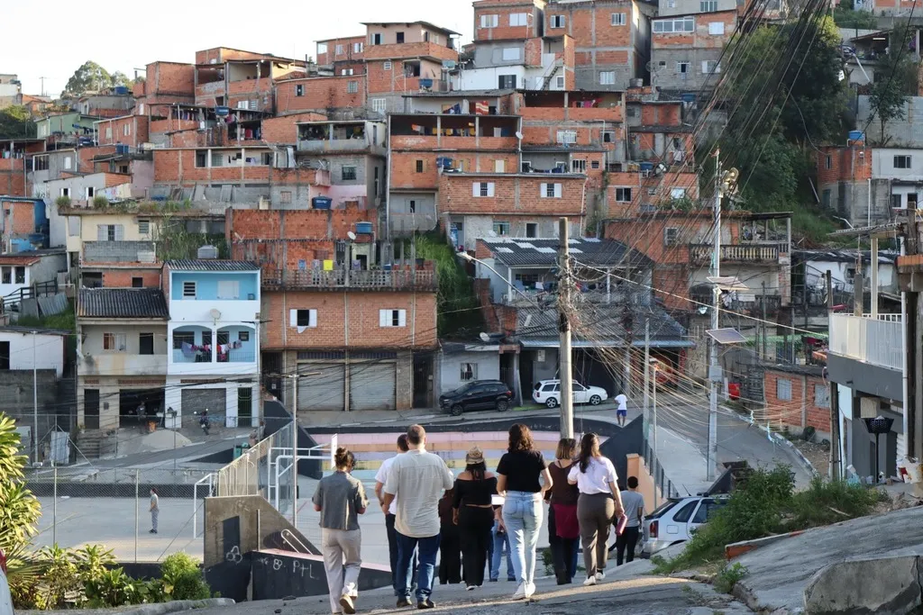 Grupo de pessoas desce uma rua íngreme, com quadra esportiva à esquerda e casas de alvenaria próximas umas das outras na encosta ao fundo. Postes e fios atravessam a paisagem